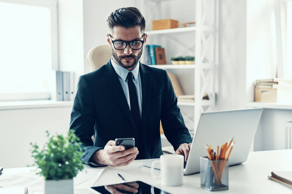Jeune homme concentré en costume d'affaires élégant travaillant en utilisant un téléphone intelligent tout en étant assis dans le bureau
 - Photo, image