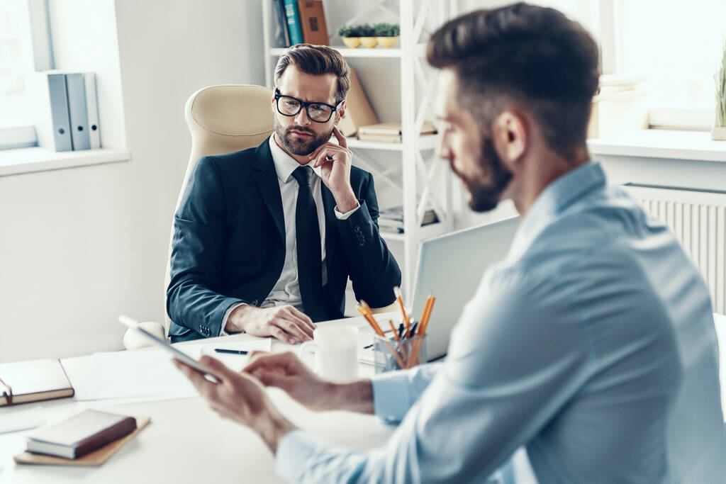 Zwei gut aussehende junge Männer in formeller Kleidung kommunizieren miteinander, während sie im Büro sitzen - Foto, Bild
