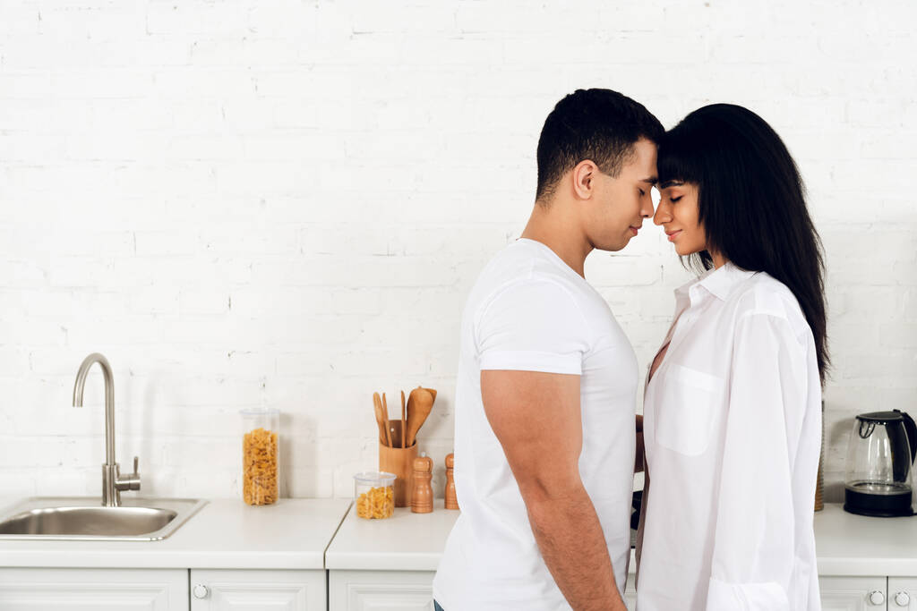 Interracial couple with closed eyes facing each other and smiling in kitchen - Photo, Image