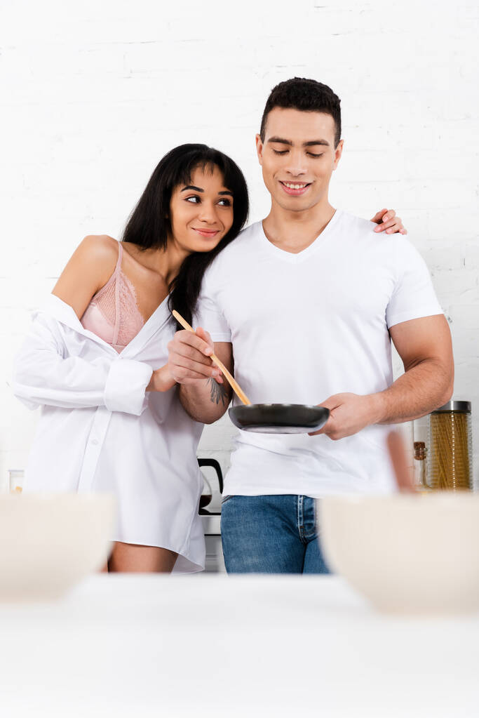 Selectieve focus van Afrikaans Amerikaans meisje glimlachende en knuffelende man met koekenpan en spatel in de keuken - Foto, afbeelding