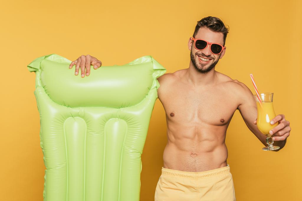 szczęśliwy człowiek bez koszuli w okularach przeciwsłonecznych trzymając kieliszek koktajlowy ze świeżym sokiem pomarańczowym i dmuchanym materacem na żółtym tle - Zdjęcie, obraz