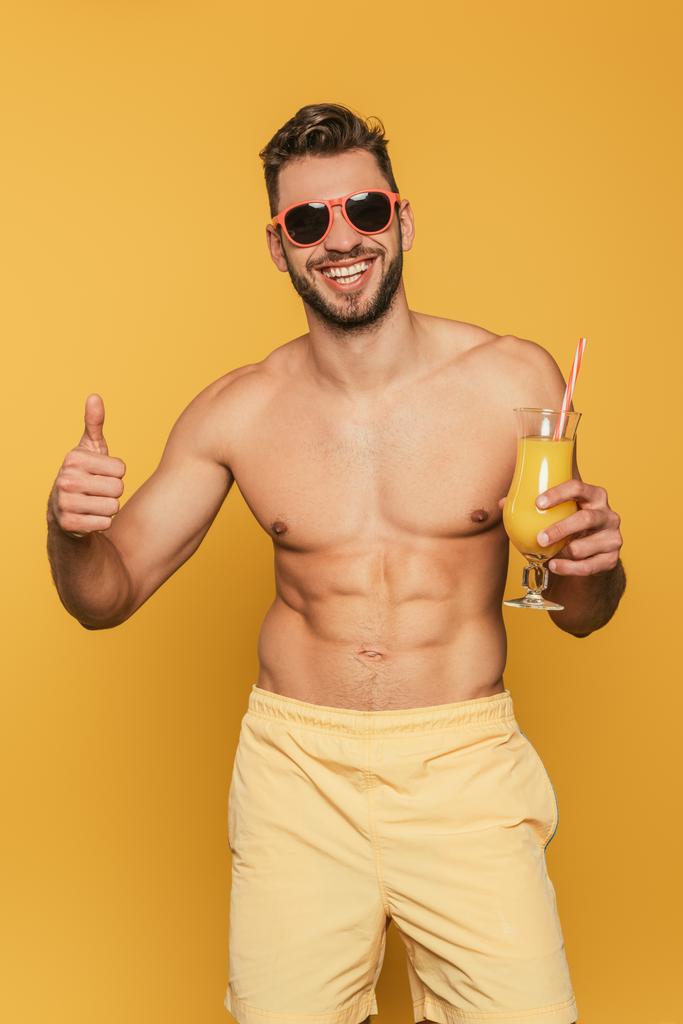 excité, homme musclé dans les lunettes de soleil montrant pouce levé tout en tenant verre à cocktail avec du jus d'orange frais sur fond jaune
 - Photo, image