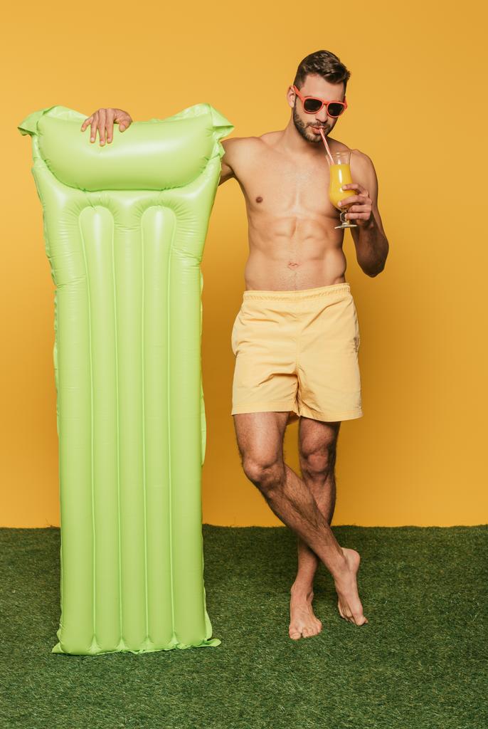 plný pohled na muže bez trička pití pomerančové šťávy a držení nafukovací matrace, zatímco stojí na zelené trávě na žlutém pozadí - Fotografie, Obrázek