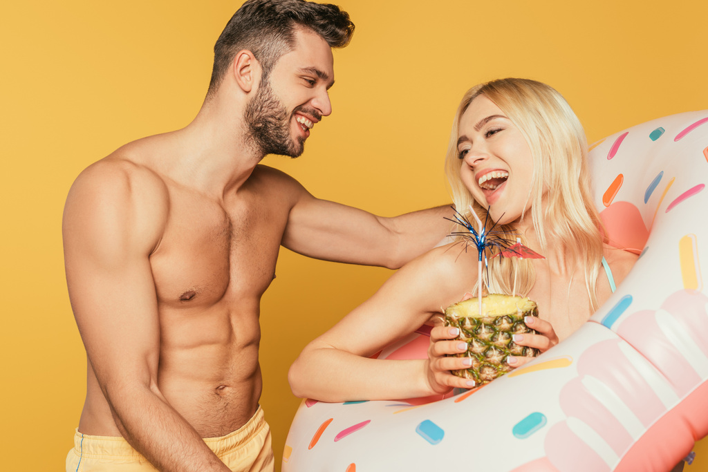 smiling muscular man touching excited girl in swim ring holding pineapple with cocktail isolated on yellow - Photo, Image