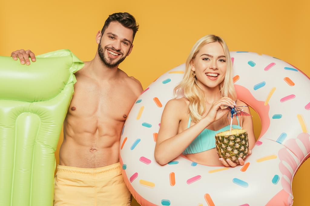 guapo camisa hombre sosteniendo colchón inflable cerca atractiva chica rubia con anillo de natación y piña con cóctel aislado en amarillo
 - Foto, Imagen