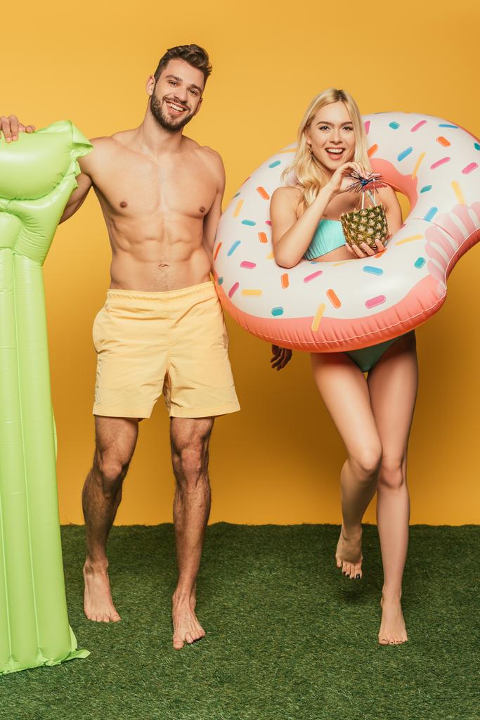 jóképű izmos férfi felfújható matrac és mosolygós szőke lány úszógyűrű és ananász koktéllal sárga alapon - Fotó, kép