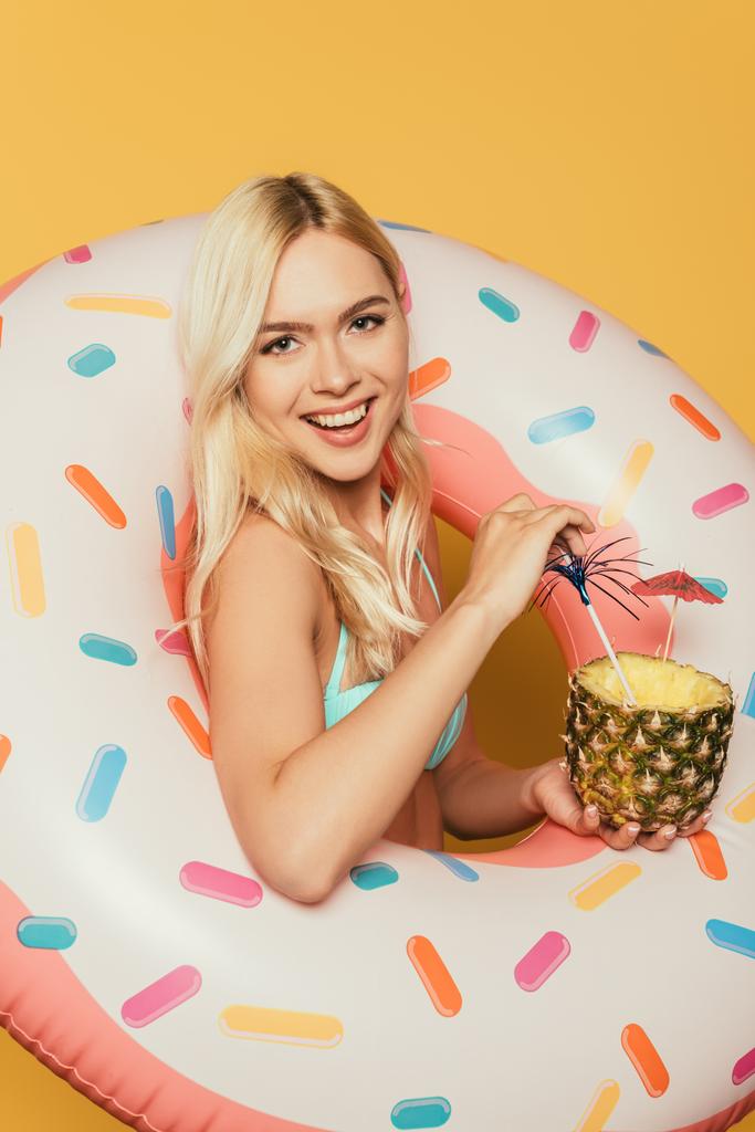 glückliches blondes Mädchen im Schwimmring hält die Hälfte der Ananas mit frischem Cocktail auf gelbem Hintergrund - Foto, Bild
