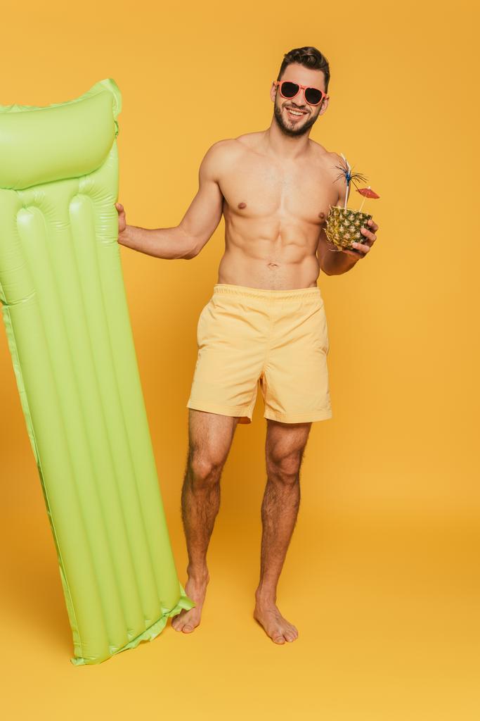 Şişme şilte tutan neşeli kaslı adam ve sarı arka planda taze kokteyl olan yarım ananas. - Fotoğraf, Görsel