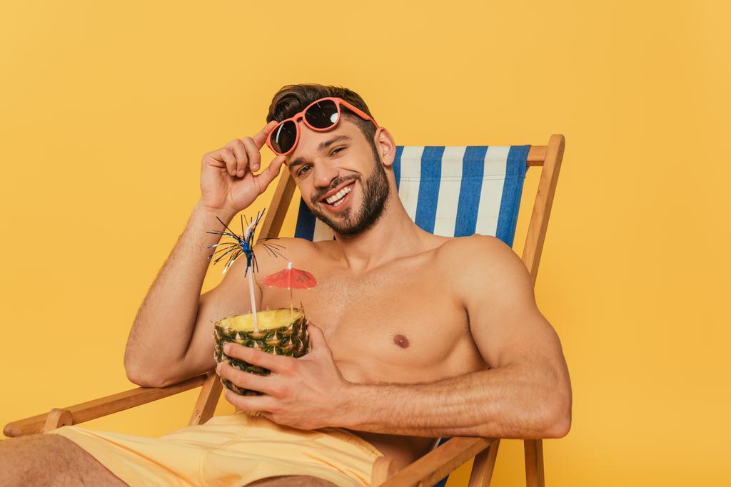feliz hombre sin camisa tocando gafas de sol y mirando a la cámara mientras sostiene la mitad de la piña con cóctel fresco aislado en amarillo
 - Foto, imagen