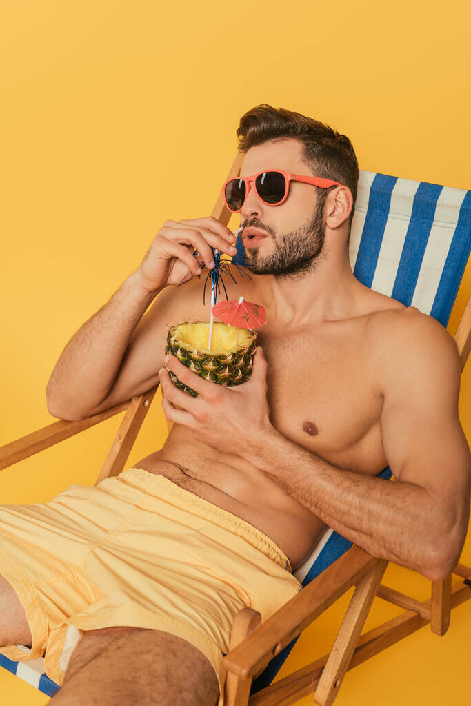 Мужчина без рубашки в солнечных очках пьет свежий коктейль из половины ананаса, расслабляясь в шезлонге, изолированном на желтом
 - Фото, изображение