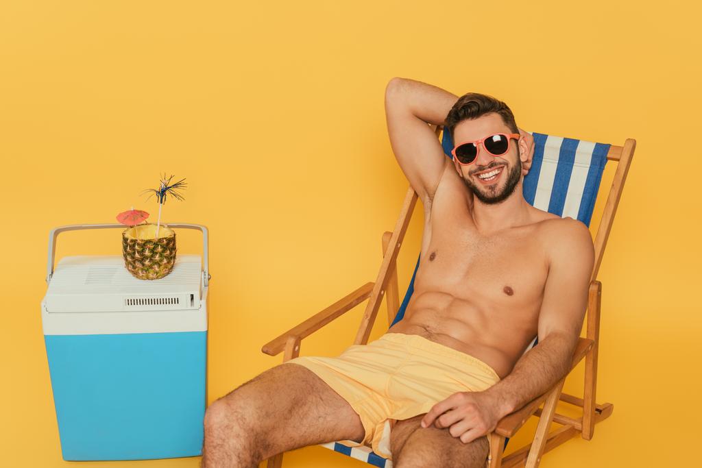 gelukkig shirtloze man in zonnebril rustend in ligstoelen in de buurt van draagbare koelkast en de helft van ananas met verse cocktail op gele achtergrond - Foto, afbeelding