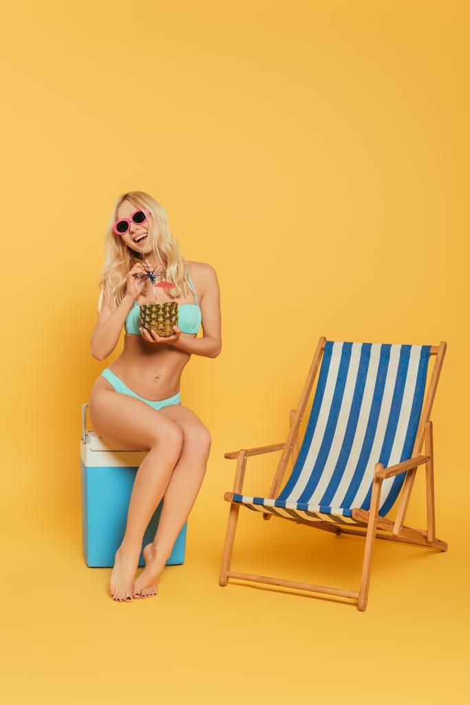 wesoła blondynka ze świeżym koktajlem w ananasie siedząca na przenośnej lodówce koło leżaka na żółtym tle - Zdjęcie, obraz