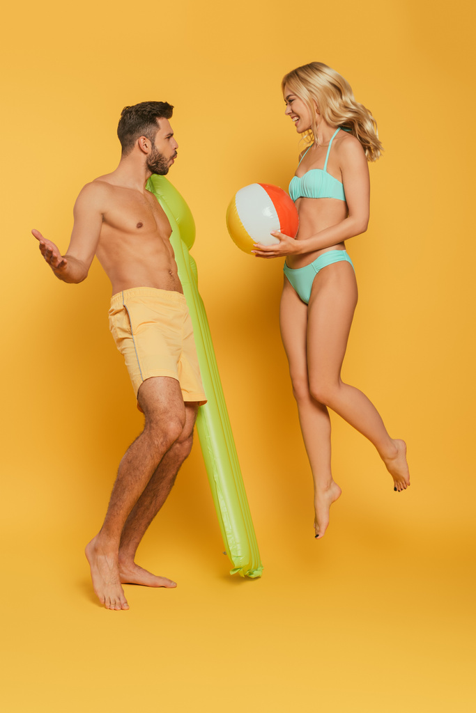 веселая девушка прыгает с надувным мячом рядом взволнованный мужчина держа надувной матрас на желтом фоне
 - Фото, изображение