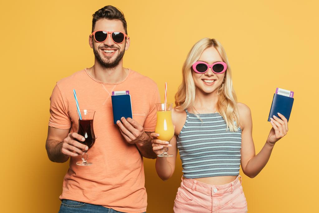 wesoła młoda para w okularach przeciwsłonecznych trzymająca koktajle i dokumenty uśmiechając się do kamery na żółtym tle - Zdjęcie, obraz