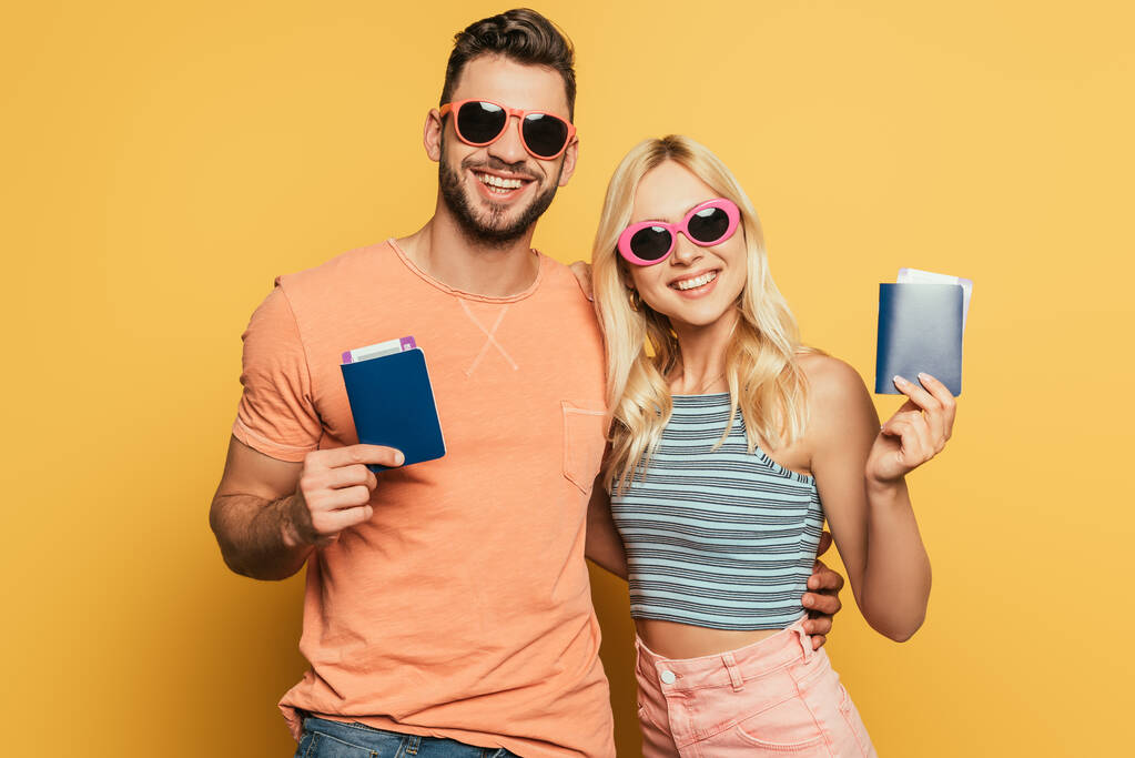 šťastný mladý pár v slunečních brýlích ukazující pasy a letenky na žlutém pozadí - Fotografie, Obrázek