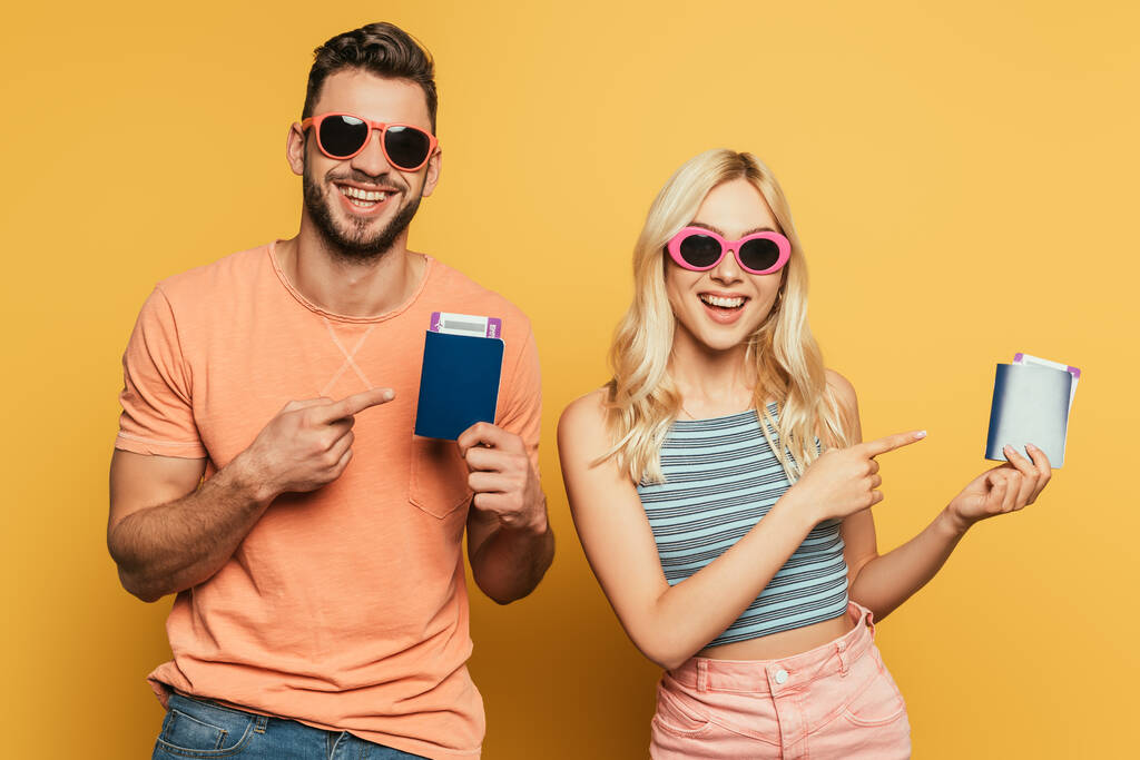 szczęśliwa młoda para w okularach przeciwsłonecznych wskazująca palcami na paszporty i bilety lotnicze na żółtym tle - Zdjęcie, obraz