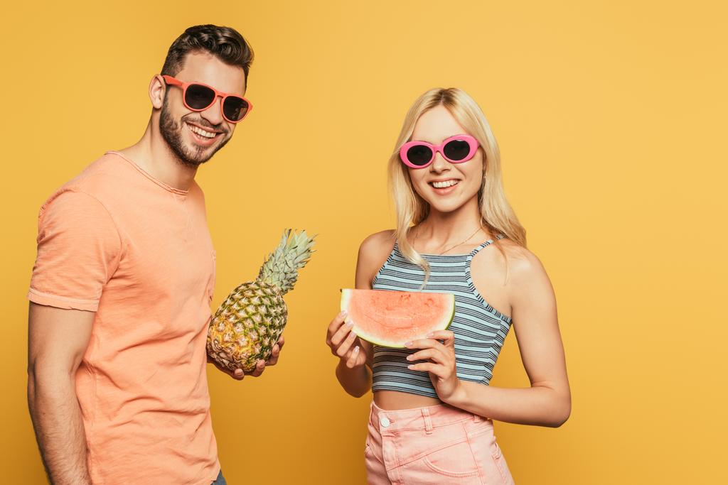 fröhlicher Mann hält ganze Ananas in der Nähe lächelndes blondes Mädchen mit Stück Wassermelone auf gelbem Hintergrund - Foto, Bild