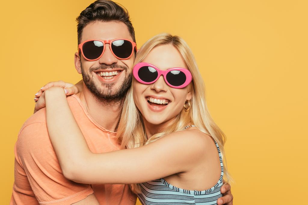 счастливый красивый мужчина и привлекательная блондинка в солнцезащитных очках обнимая изолированные на желтый
 - Фото, изображение