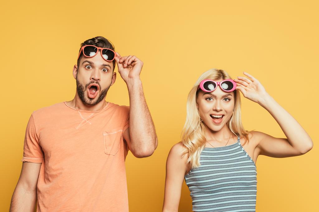 shocked man and woman touching sunglasses while looking at camera on yellow background - Photo, Image