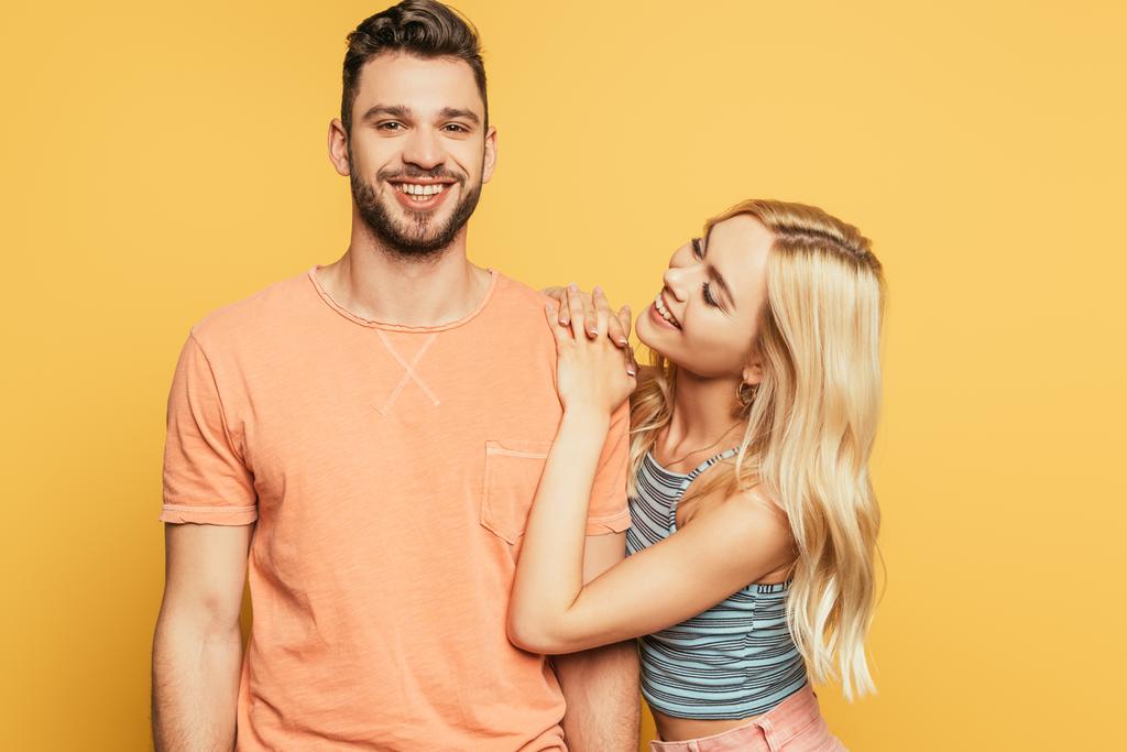 happy blonde girl embracing shoulder of smiling man isolated on yellow - Photo, Image
