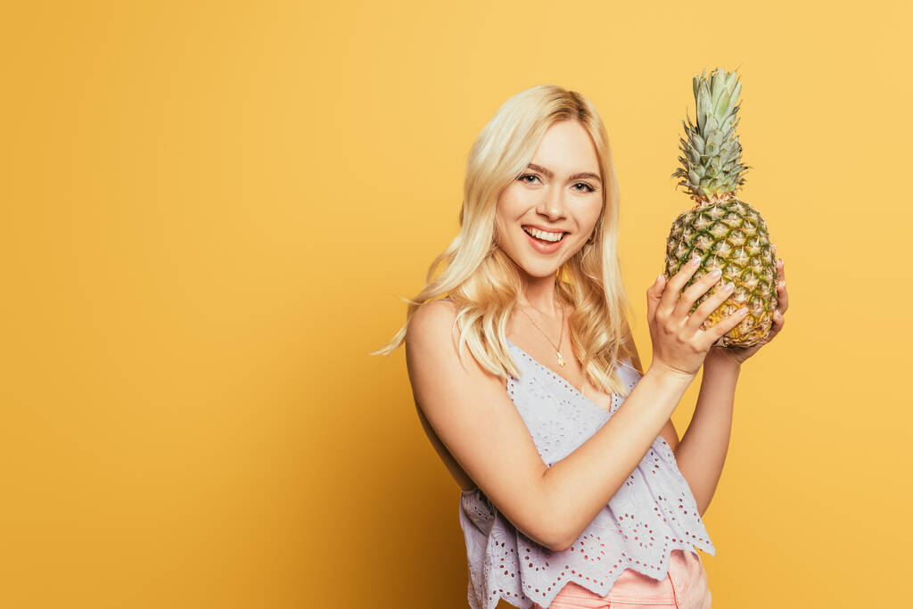 glückliches blondes Mädchen hält Ananas in der Hand und lächelt in die Kamera auf gelbem Hintergrund - Foto, Bild