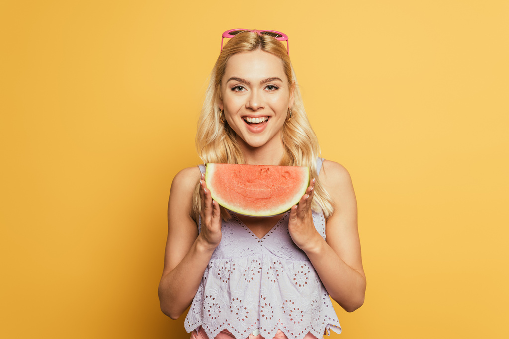 houkutteleva blondi tyttö tilalla siivu vesimelonia ja hymyilee kameralle keltaisella taustalla
 - Valokuva, kuva