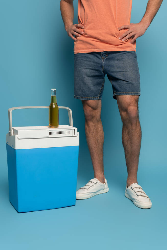 vista recortada del hombre de pie con las manos en las caderas cerca de la botella de cerveza en la nevera portátil sobre fondo azul
 - Foto, Imagen