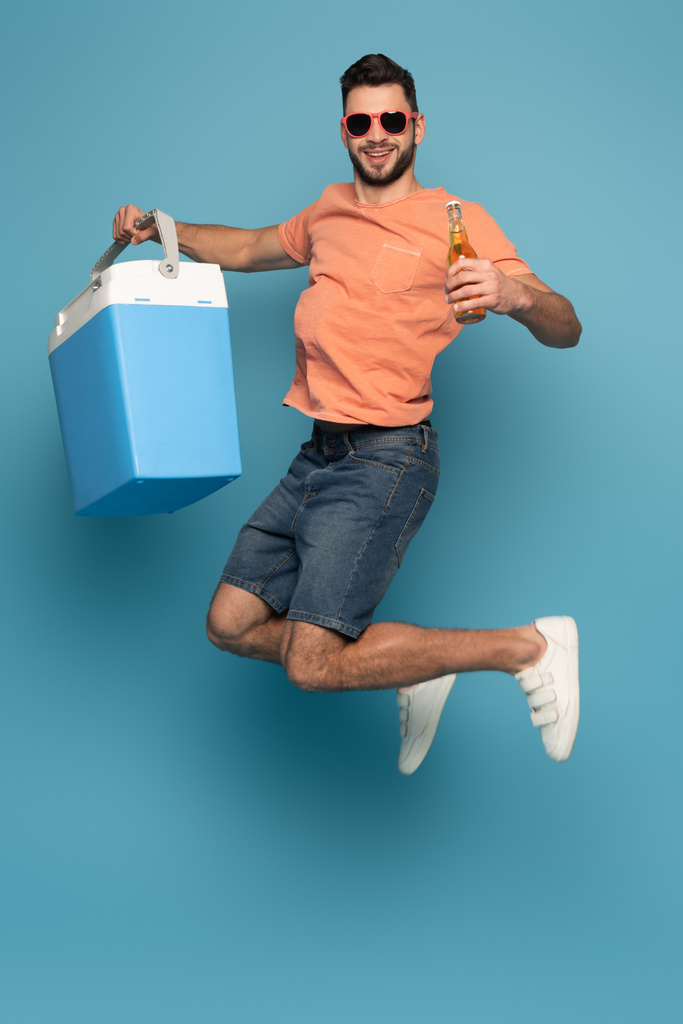 ενθουσιασμένοι άνθρωπος σε γυαλιά ηλίου άλμα, ενώ κρατώντας μπουκάλι μπύρα και φορητό ψυγείο σε μπλε φόντο - Φωτογραφία, εικόνα