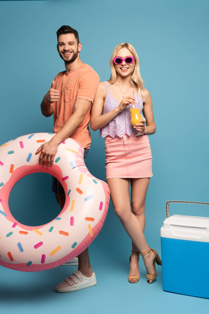 hombre feliz con anillo de natación que muestra el pulgar hacia arriba, y la niña sonriente sosteniendo un vaso de jugo de naranja cerca de la nevera portátil sobre fondo azul
 - Foto, Imagen