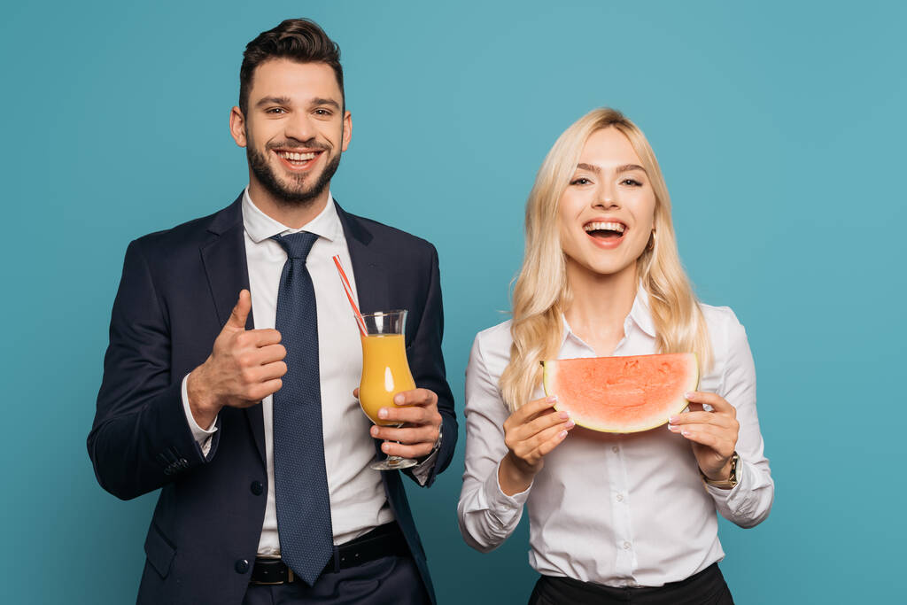 szczęśliwy biznesmen z sokiem pomarańczowym pokazując huk w górę w pobliżu uśmiechnięty bizneswoman z soczysty arbuz na niebieskim tle - Zdjęcie, obraz