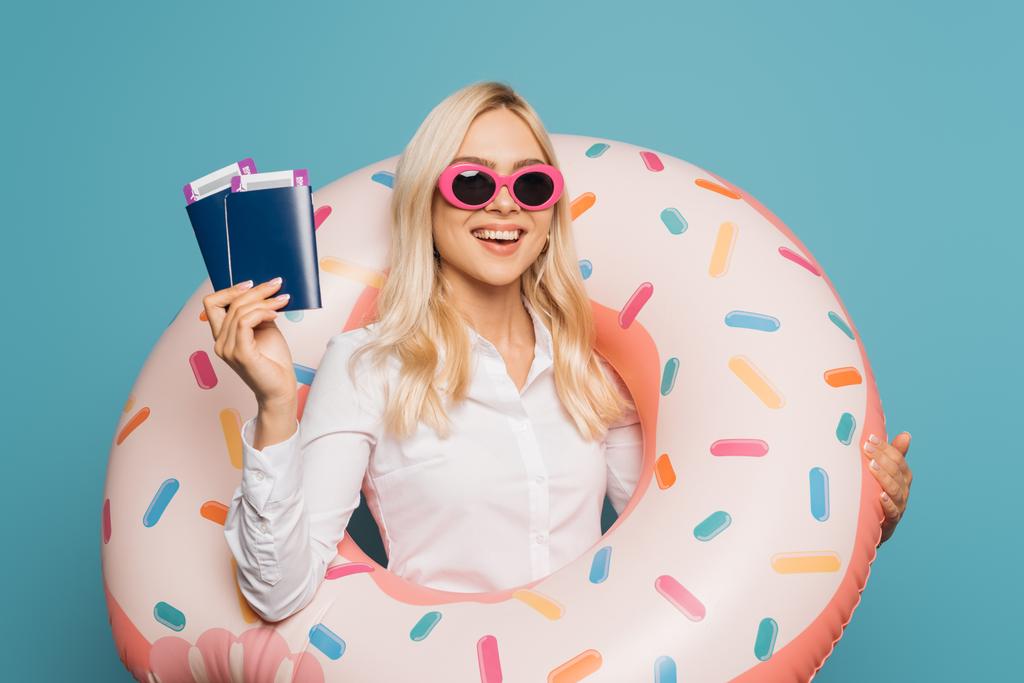 femme d'affaires heureuse en lunettes de soleil et anneau de natation tenant passeports et billets d'avion isolés sur bleu
 - Photo, image