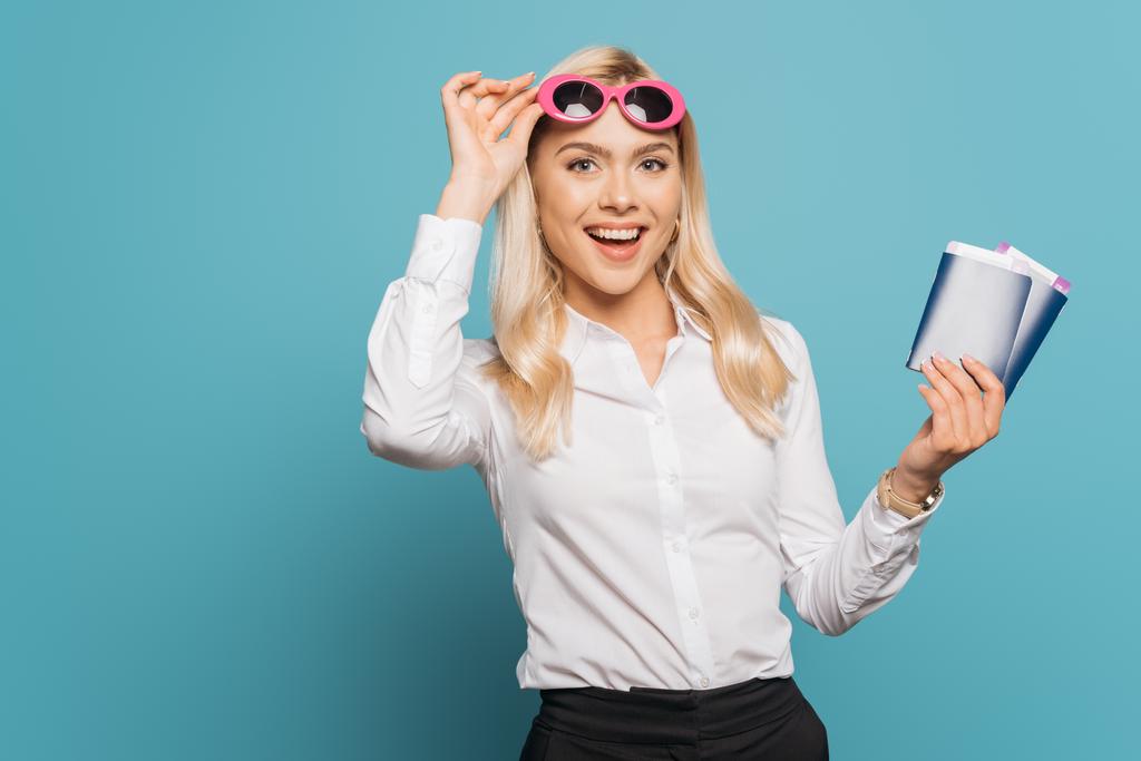 glückliche Geschäftsfrau mit Sonnenbrille, während sie Reisepässe und Flugtickets auf blauem Hintergrund hält - Foto, Bild