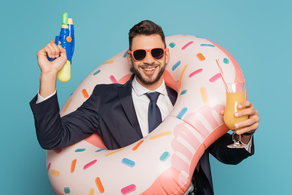 alegre hombre de negocios en anillo de natación sosteniendo pistola de agua y vaso de jugo de naranja sobre fondo azul
 - Foto, imagen