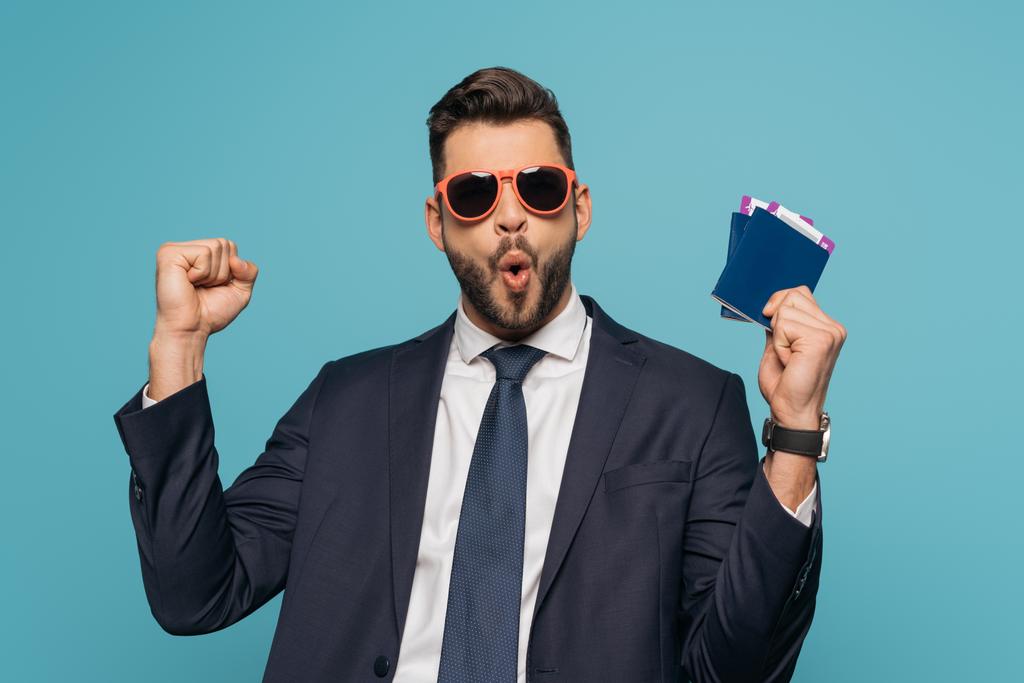 возбужденный бизнесмен в солнечных очках показывает жест победителя, держа в руках паспорта и авиабилеты изолированы на синий
 - Фото, изображение