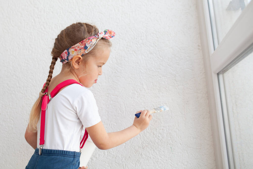 Kleine hübsche Mädchen malt eine weiße Wand mit einem Pinsel, ein Kind und Reparaturen in der Wohnung, drinnen - Foto, Bild