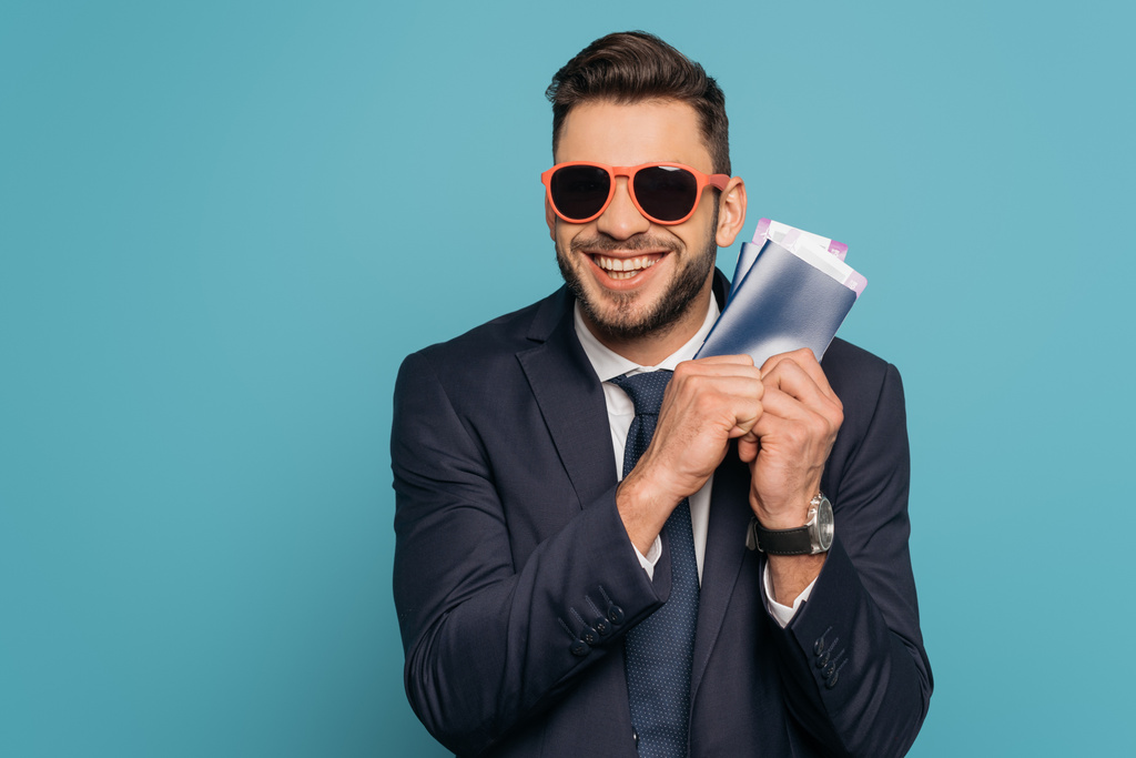 excitado hombre de negocios en gafas de sol con pasaportes y billetes de avión mientras sonríe a la cámara aislada en azul
 - Foto, imagen