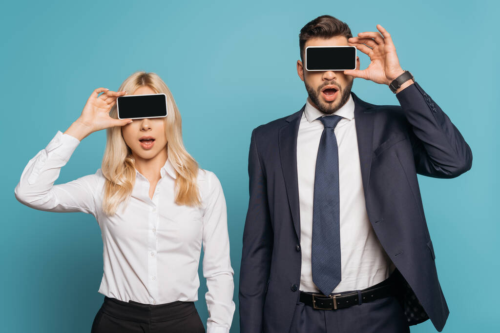 sorprendido hombre de negocios y mujer de negocios cubriendo los ojos con teléfonos inteligentes con pantalla en blanco sobre fondo azul
 - Foto, Imagen