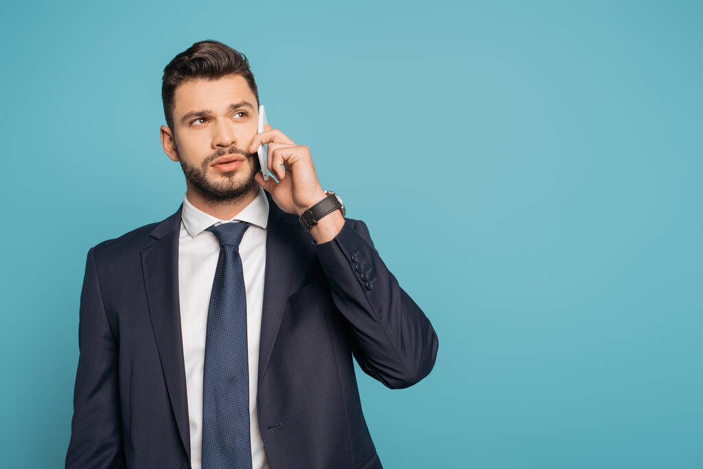 serious, handsome businessman talking on smartphone and looking away isolated on blue - Photo, Image