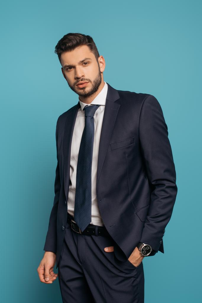 serious, handsome businessman looking at camera while holding hand in pocket on blue background - Photo, Image
