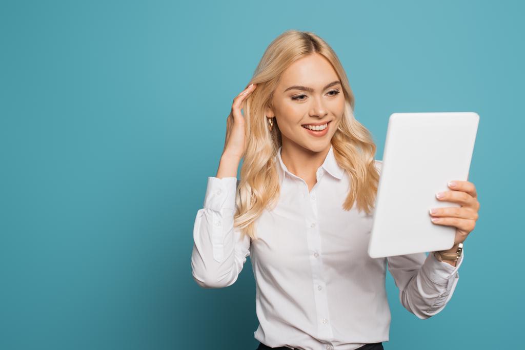 attraktive, blonde Geschäftsfrau, die Haare berührt und lächelt, während sie ein digitales Tablet auf blauem Hintergrund benutzt - Foto, Bild