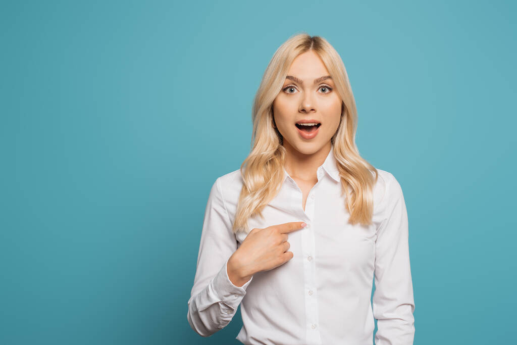 surprised businesswoman pointing with finger at herself while looking at camera on blue background - Photo, Image
