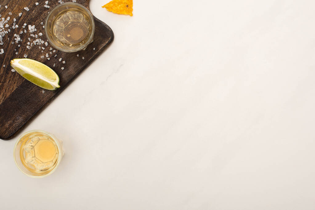 vrchní pohled na zlatou tequilu s vápnem, sůl na dřevěné řezací desce na bílém mramorovém povrchu - Fotografie, Obrázek