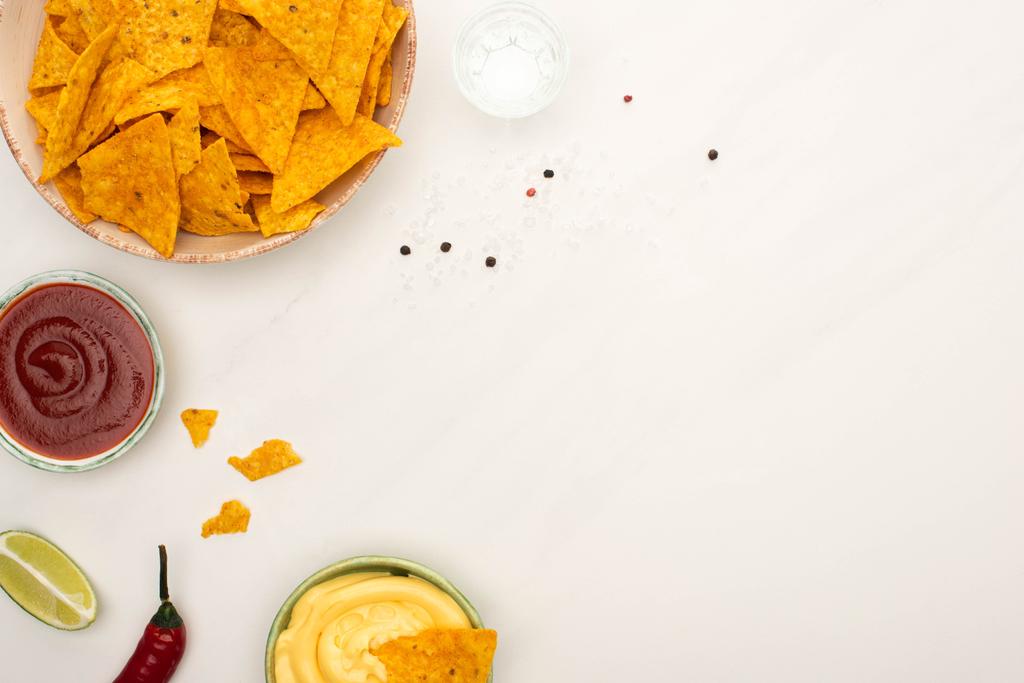 bovenaanzicht van tequila met limoen, chili peper, sauzen en nacho 's op wit marmeren oppervlak - Foto, afbeelding