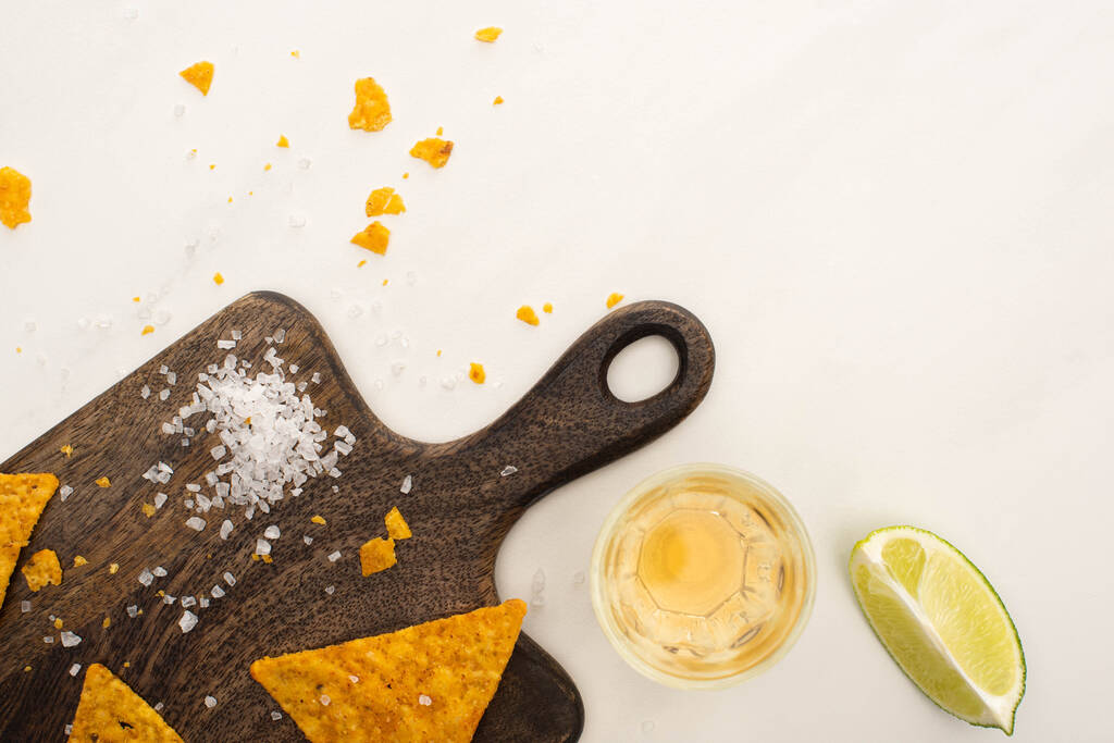 top view of golden tequila with lime, salt and nachos on wooden cutting board on white marble surface - Photo, Image