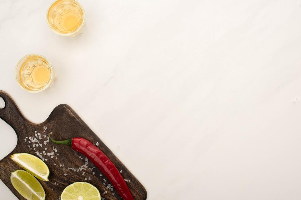 bovenaanzicht van gouden tequila met limoen, chili peper, zout op houten snijplank op wit marmeren oppervlak - Foto, afbeelding