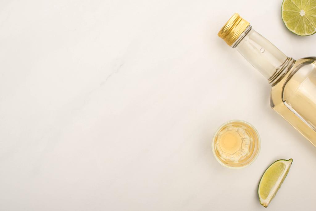 πάνω όψη της χρυσής τεκίλας σε μπουκάλι και σφηνάκι με λάιμ σε λευκή μαρμάρινη επιφάνεια - Φωτογραφία, εικόνα