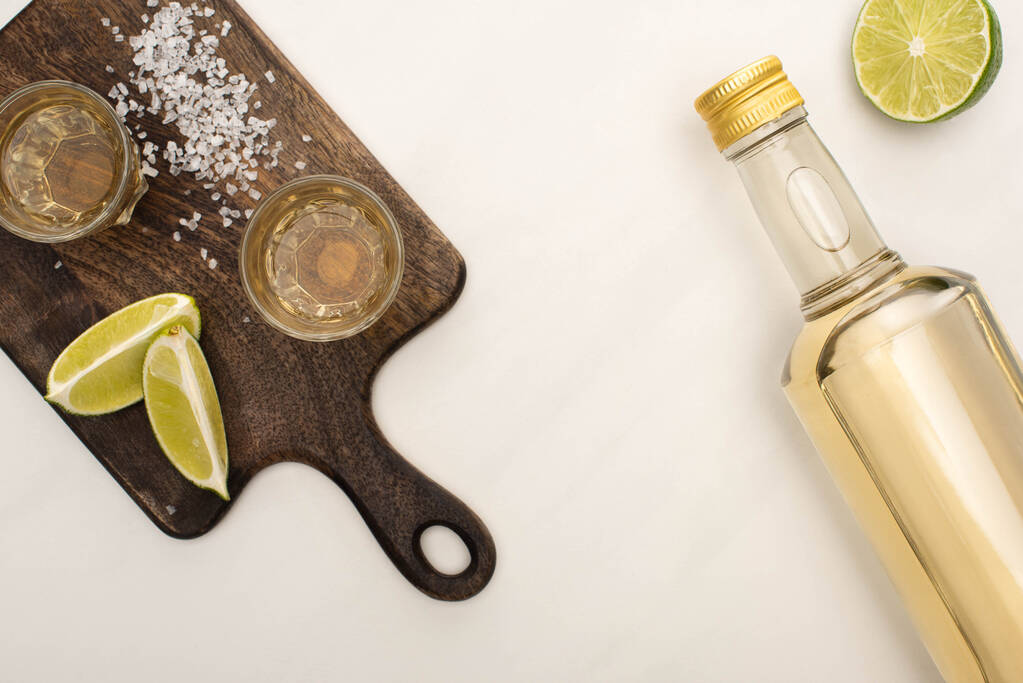 Blick von oben auf goldenen Tequila mit Kalk, Salz auf Holzschneidebrett auf weißem Marmor - Foto, Bild