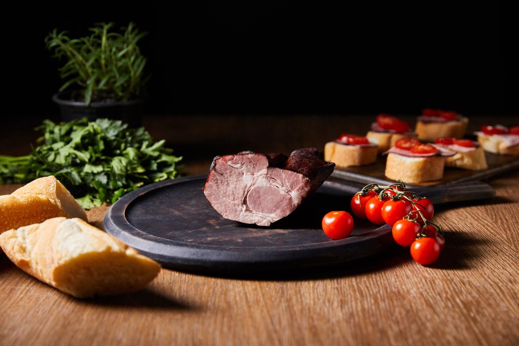 foyer sélectif de jambon savoureux à bord près de canape, persil, tomates cerises et baguette sur table en bois isolé sur noir
 - Photo, image
