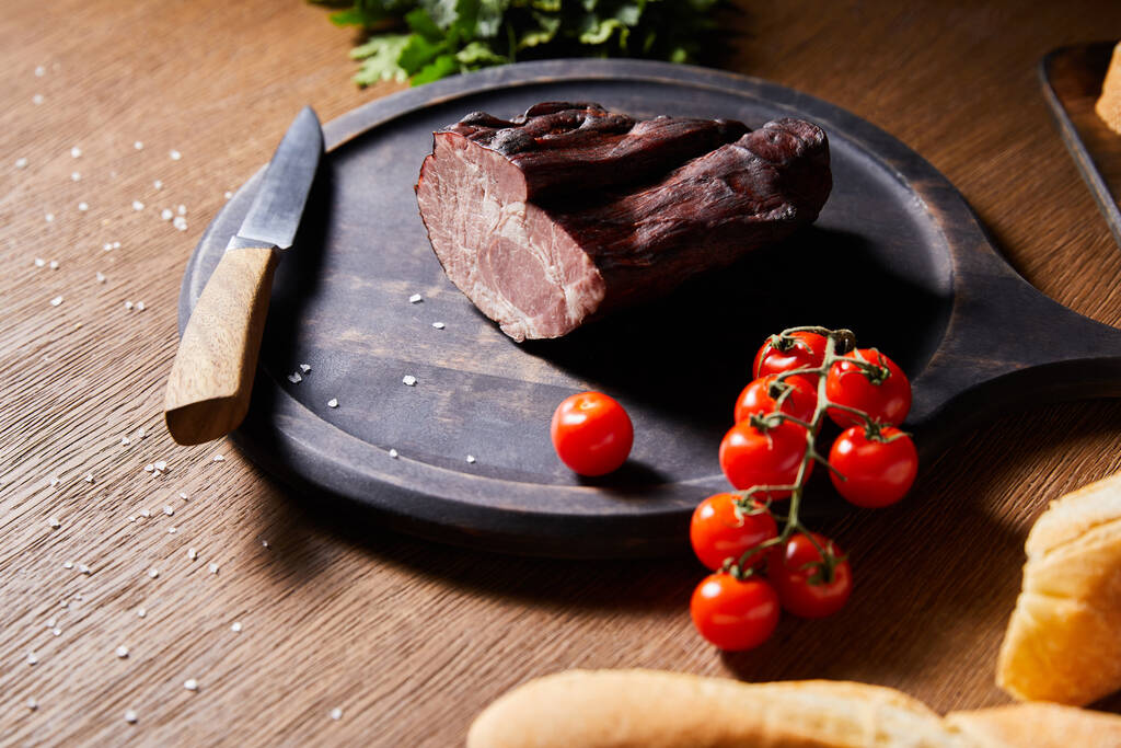 foco seletivo de presunto saboroso a bordo perto de salsa, tomates cereja e baguete com sal espalhado e faca na mesa de madeira
 - Foto, Imagem