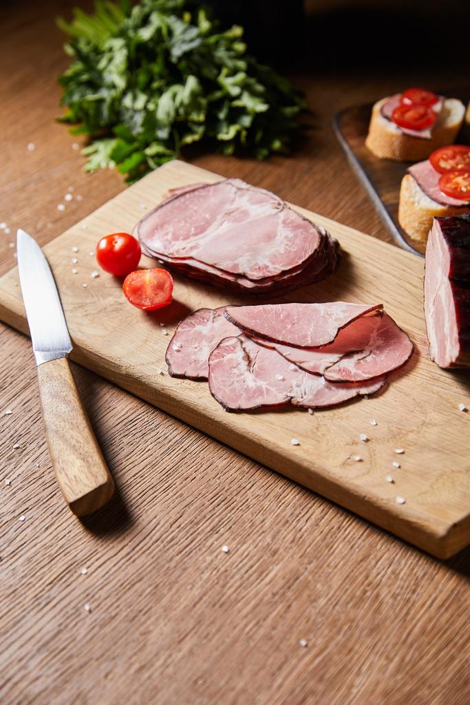 selectieve focus van smakelijke ham plakjes, kerstomaten en mes op snijplank bij peterselie en canape - Foto, afbeelding